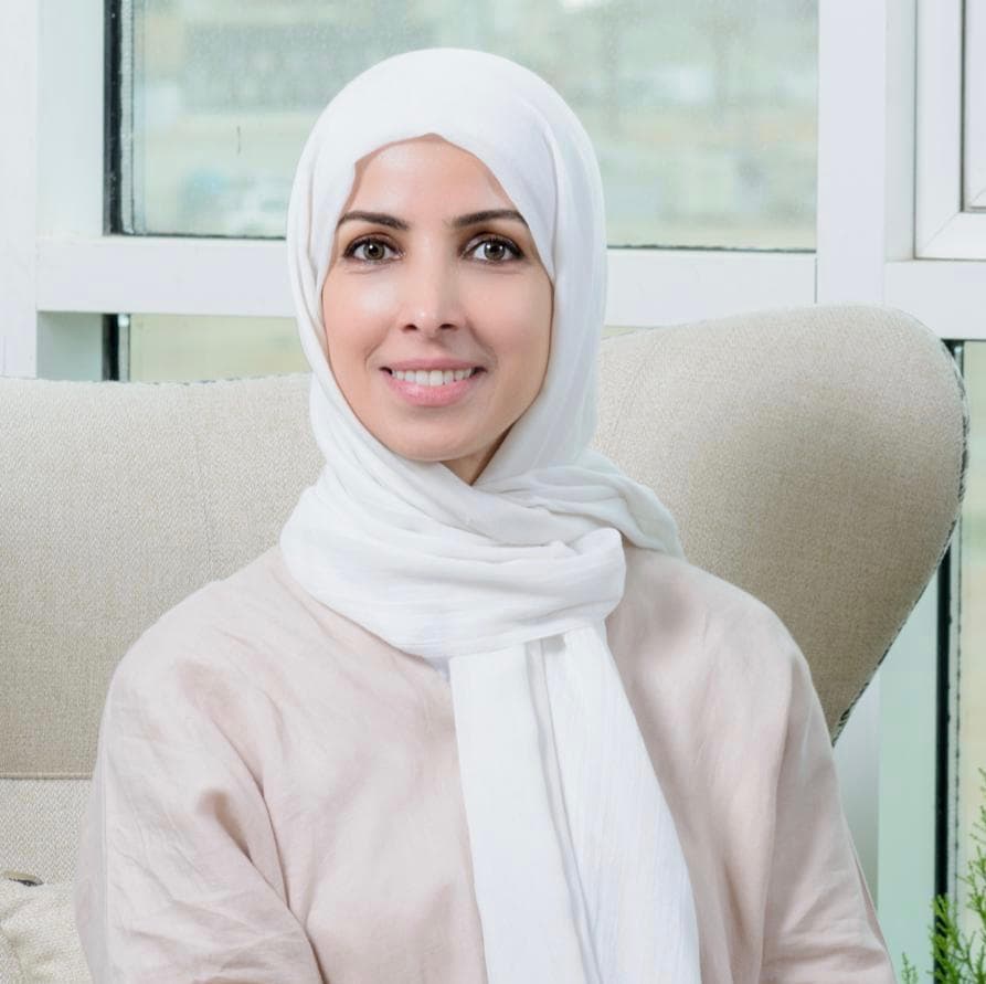 نائلة السقاف مبتسمة، ترتدي حجابًا أبيض وقميصًا بلون بيج فاتح، وتجلس على كرسي بجانب نافذة