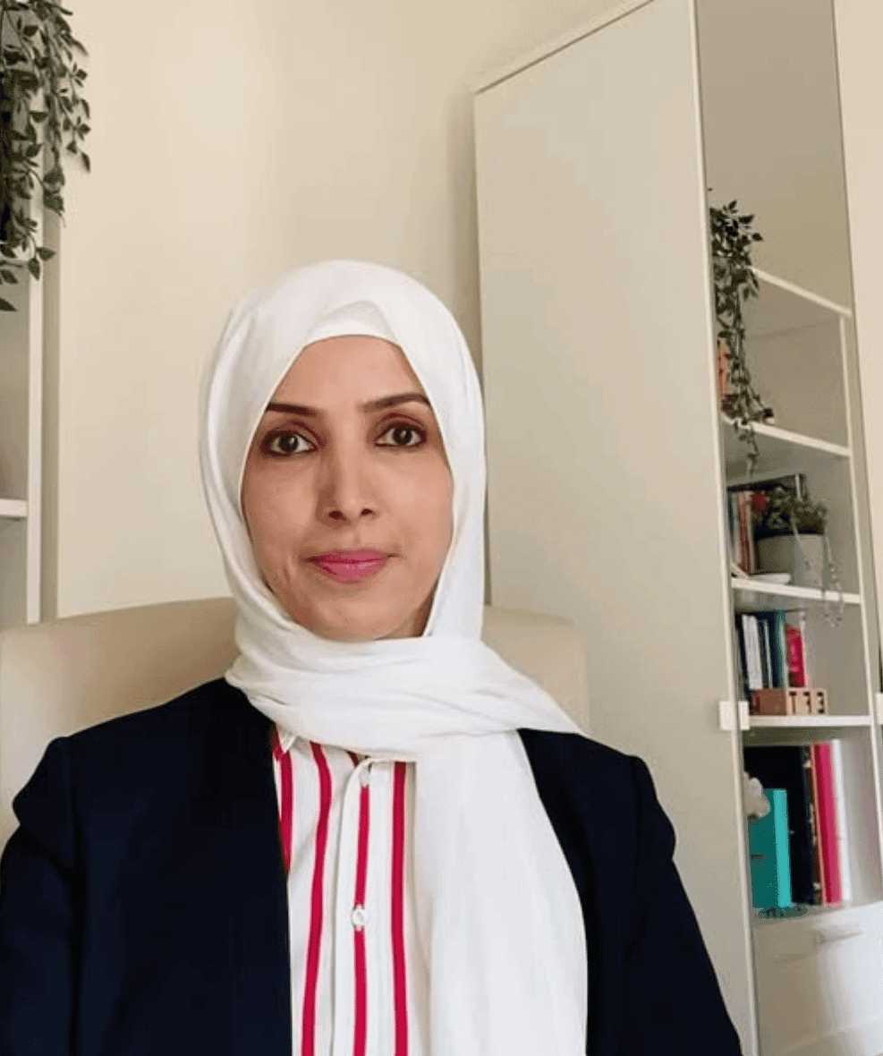 نائلة السقاف ترتدي حجابًا أبيض وسترة كحلية فوق قميص مخطط، في مكتبها أمام رفوف كتب